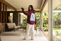 A woman stands in a modern indoor-outdoor corridor wearing a maroon hoodie with a large light blue graphic print on the back and white sweatpants, looking over her shoulder.