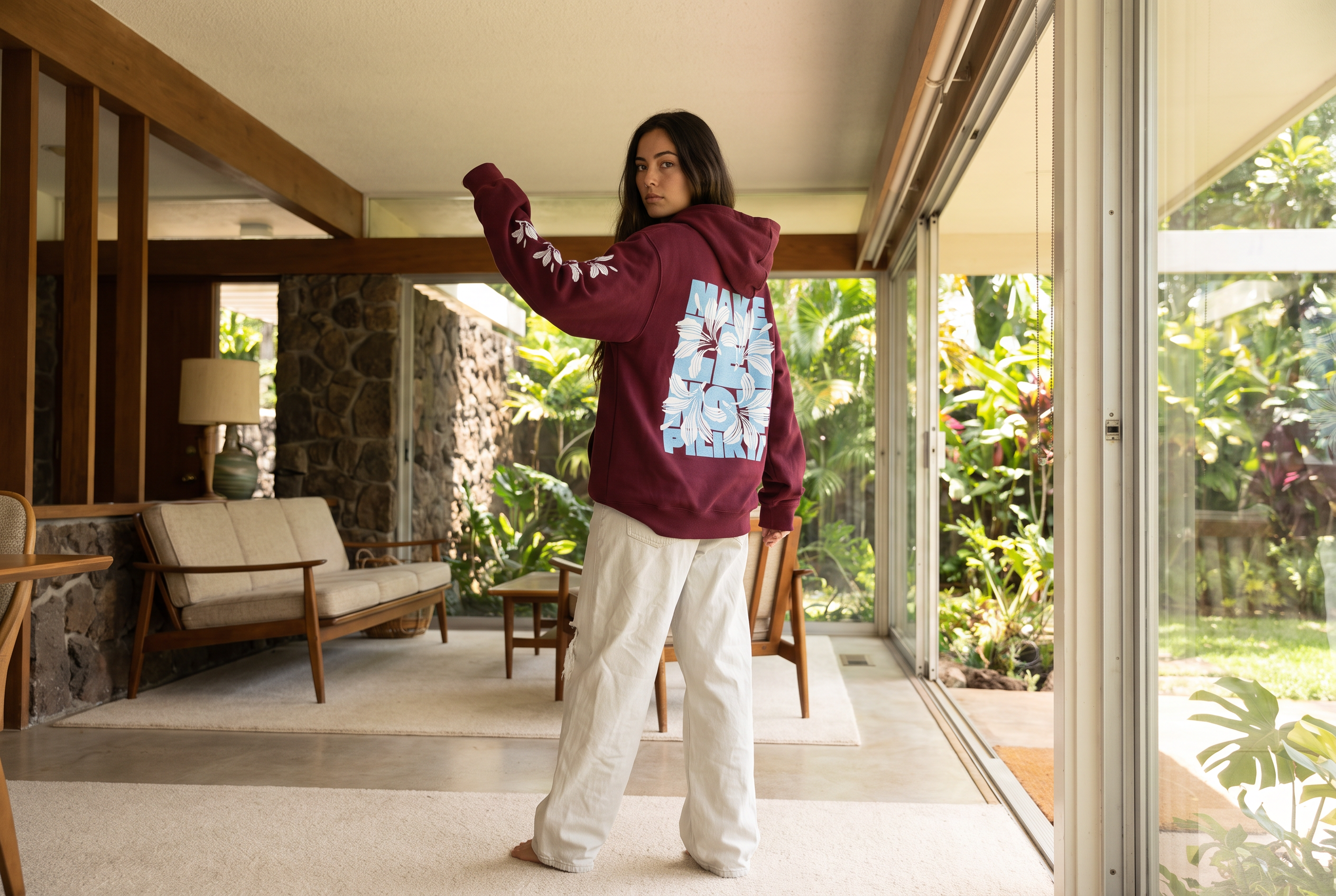 A woman stands in a modern indoor-outdoor corridor wearing a maroon hoodie with a large light blue graphic print on the back and white sweatpants, looking over her shoulder.